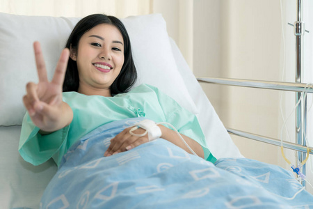 年轻的亚洲患者妇女躺在病床上, 用盐水滴注显示胜利的迹象。滴盐水液帮助病人