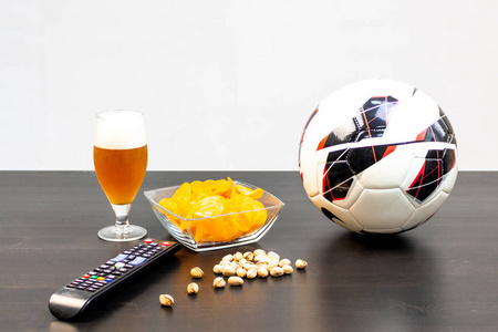 人们准备用啤酒在电视上观看足球比赛。桌子上有啤酒, 球, 电视遥控器, 小吃。工艺啤酒。光背景