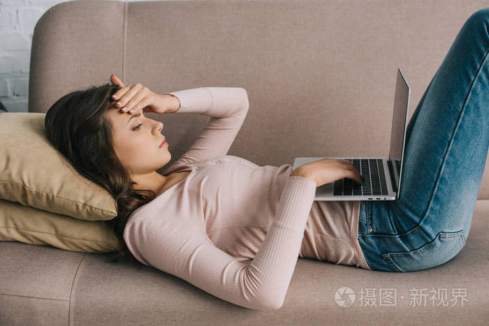 在沙发上躺着膝上型电脑的年轻女性头痛的侧面观察