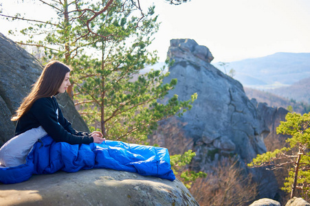 美丽的年轻长发女子独自坐在大山岩的睡袋上, 享受着晴朗的夏日清晨的美景, 旅游和积极的生活方式概念