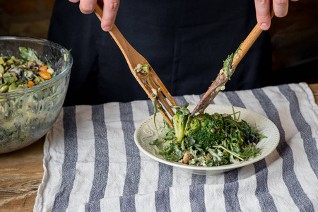 男士特写照片在餐桌上用木勺和叉子在玻璃碗里吃美味的绿色沙拉和芝麻菜和菠菜。