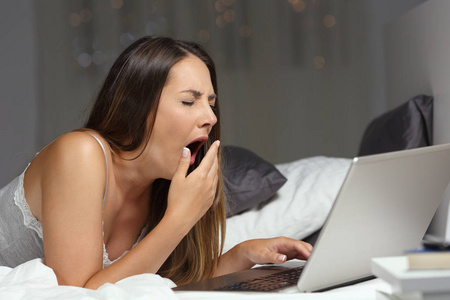 疲惫的女人在家里晚上用笔记本电脑在床上打呵欠