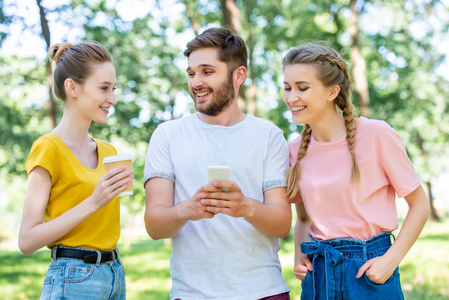 微笑的朋友与咖啡一起去使用智能手机在公园