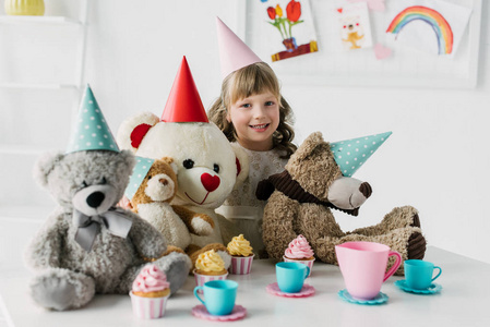 微笑的生日孩子与泰迪熊在锥体有茶党与蛋糕在桌