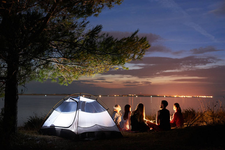 五名游客, 妇女和男子坐在海边的篝火周围的帐篷在水面上的树上, 在日落背景下的蓝色傍晚的天空。旅游业和野营概念