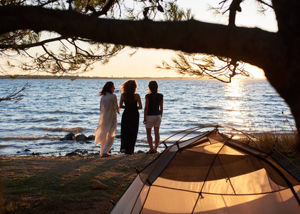 三名年轻女子坐在湖岸边, 在旅游帐篷前的树下, 在清澈的水背景下享受美丽的日落。旅游, 友谊和露营概念