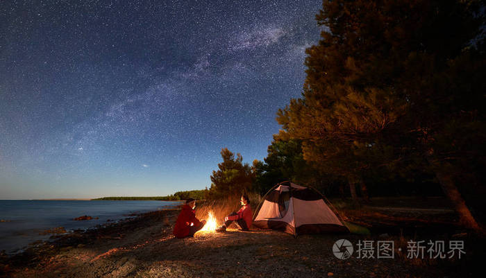 年轻男女游客坐在帐篷前的篝火旁 在蓝色的水和森林背景下 在星光