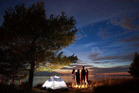 四名游客的后景色站在海边的篝火旁, 在蓝色傍晚的天空下, 夕阳下的第一颗星星在帐篷附近。旅游友谊野营与自然美景
