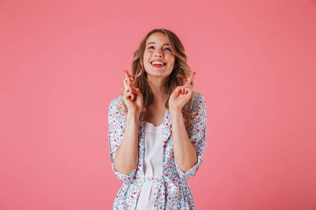 一个兴奋的年轻女子在夏季礼服的肖像握住手指交叉的好运, 并期待在粉红色背景孤立