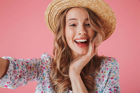 关闭一个开朗的年轻女子的肖像在夏季礼服和草帽采取自拍孤立的粉红色背景