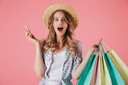 一个惊讶的年轻女子在夏季礼服和草帽的肖像在手持购物袋, 在粉红色的背景下, 用手机交谈