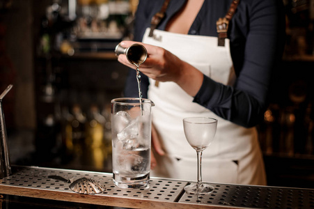 女酒保在白色围裙增加酒精入玻璃与冰块在酒吧柜台