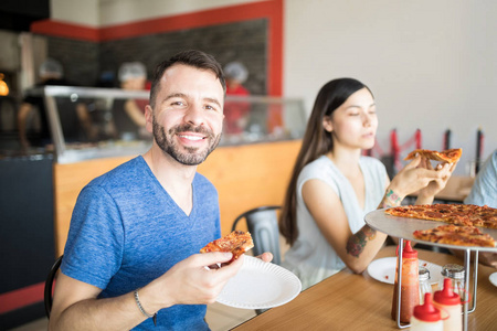 年轻英俊男子的肖像手持香肠比萨饼片在手坐在餐厅与朋友在后台, 而看着相机