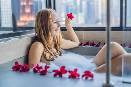 年轻女子坐在浴缸与泡沫和花式花朵反对全景窗口的背景俯瞰摩天大楼和大晚上城市