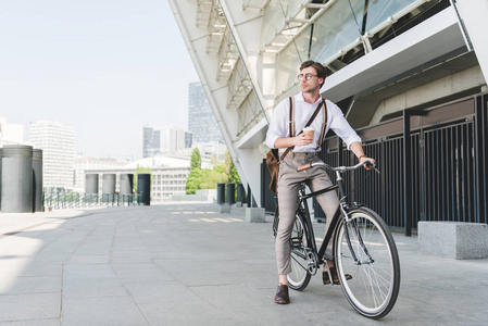 英俊的年轻人与纸杯咖啡在老式自行车附近体育体育场大厦
