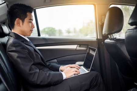英俊的年轻商人使用笔记本电脑坐在后座汽车