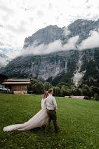 年轻的新娘和新郎走在绿色的山草甸与云在阿尔卑斯