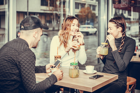 欢快的一群朋友, 一个男人和两个女人坐在桌边的咖啡厅里, 聊着开心的笑着大笑起来。女孩喝柠檬水