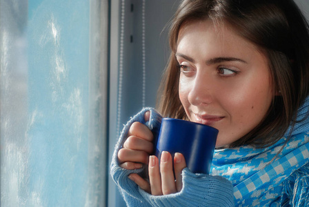 在冬天的冻结的窗口附近的杯子在毛线衣的共同的微笑的梦想女孩