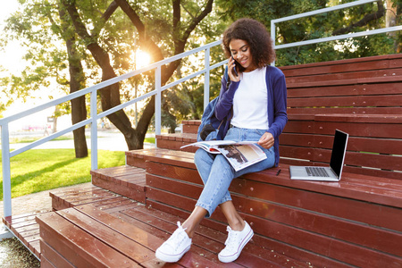 一个微笑的年轻非洲女孩的肖像与背包在公园休息时谈论手机, 阅读杂志