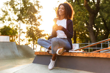 一个微笑的年轻非洲女孩的肖像与背包使用手机, 而在公园休息