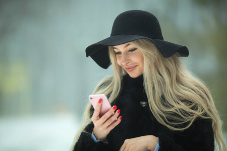 美丽的年轻女孩在毛皮大衣和帽子漫步在公园冬天的天气与电话在手
