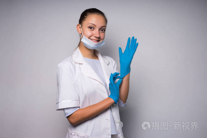年轻的微笑医生女孩穿着白色的医疗礼服穿上蓝色的手套, 看着相机