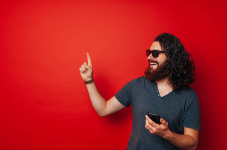 微笑幸福胡子的人手持智能手机, 并指出在 copyspace 在红色背景