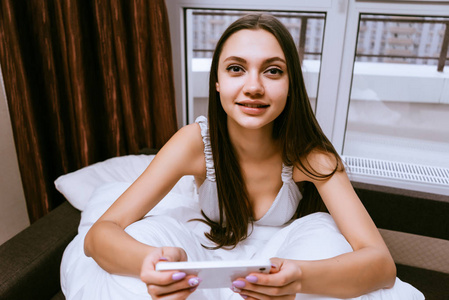 可爱的年轻女孩坐在床下的白色毯子, 手持智能手机