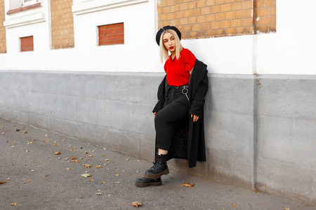 时髦美丽的年轻女子在黑色大衣与红色 tshirt 和贝雷帽附近的建筑物