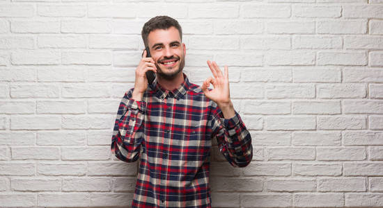 年轻的成年男子说话的电话站在白色砖墙做 ok 符号与手指, 优秀的符号