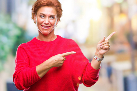 活跃的高级高加索红发妇女穿着冬季毛衣在孤立的背景微笑着, 看着相机指向镜头, 用两只手和手指指着一边