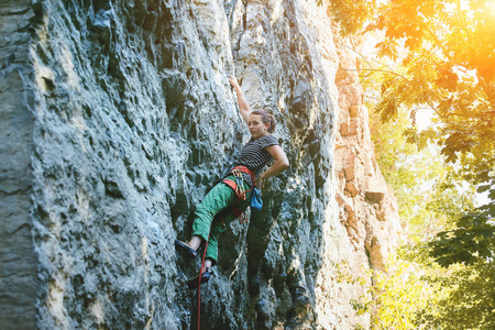 女攀岩者的侧面看着明亮的绿色裤子爬上悬崖。攀岩者爬上一堵石墙。妇女做坚硬行动