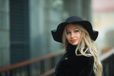 年轻美丽的女孩在黑色毛皮大衣和帽子在街道附近的建筑物