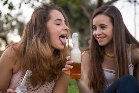 两个年轻的笑女人摆在公园的草坪上, 喝啤酒的乐趣