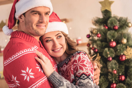 丈夫和妻子在圣诞老人的帽子拥抱附近的圣诞树在家里和看远
