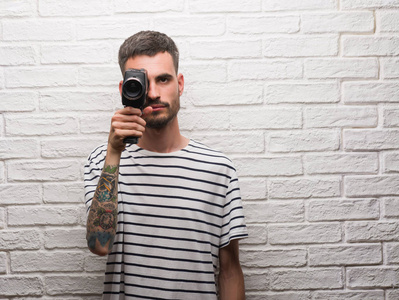 年轻人拍摄手持摄像机站在白色砖墙上, 自信的表情对聪明的面孔认真思考