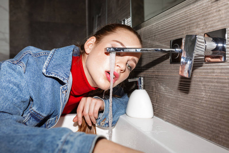 年轻漂亮女人的肖像牛仔夹克饮用自来水从水槽隔离
