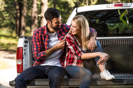 微笑的年轻夫妇拥抱和坐在汽车树干室外