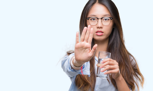 年轻的亚洲妇女喝一杯水在孤立的背景下, 张开手做停止标志认真和自信的表达, 防御姿态