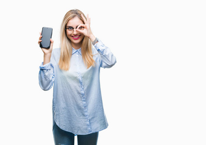 年轻美丽的金发女商人显示屏幕上的智能手机在孤立的背景与快乐的脸微笑做确定的标志与眼睛看着通过手指