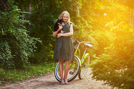 全身肖像的一个微笑的小金发女郎在休闲礼服, 持有可爱的斯皮茨狗。在公园骑自行车