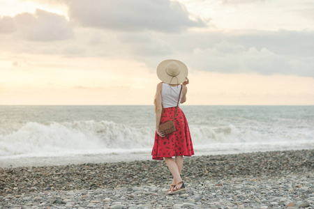 女孩在红色裙子和帽子站立在海, 日落。后退视图