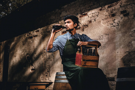 裔酿酒大师坐在木桶上, 捧着一杯工艺啤酒, 下班后放松。