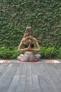 女练瑜珈的后观莲姿在墙面覆盖着绿叶的背后做着合十手势