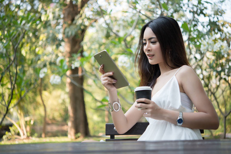 年轻美丽的女人持有一次性咖啡杯, 而短信通过智能手机在大自然户外