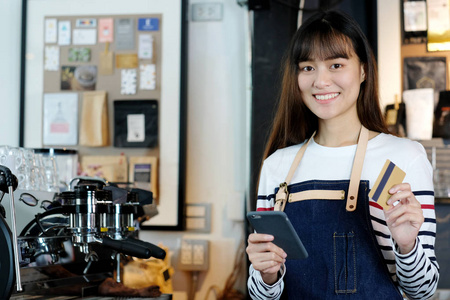 年轻的亚洲妇女咖啡师使用智能手机和持有信用 ca