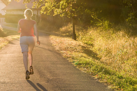 运动妇女在乡间小路上奔跑在日出的灯光下。美丽的女性跑户外在绿叶和绿色郊区