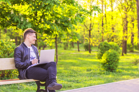 一个年轻英俊的男人坐在公园里用笔记本电脑工作。那个自由职业者在外面工作, 微笑着