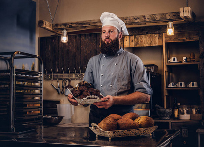 穿着制服的英俊的大胡子厨师在面包店厨房里展示新鲜面包托盘